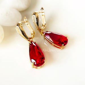 May include: A pair of gold-colored drop earrings with large, teardrop-shaped, red gemstones. The earrings have a lever-back closure and are displayed on a white surface. The earrings are a type of jewelry.