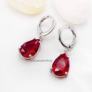 May include: A pair of silver-toned drop earrings with teardrop-shaped, deep red gemstones. The earrings have a lever-back closure and are displayed on a white surface with a few decorative objects.