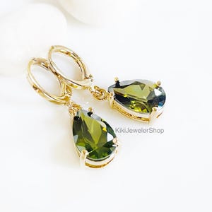May include: Gold-tone drop earrings with olive green pear-shaped gemstones. The earrings feature a lever back closure and are displayed on a white surface. The gemstones have a faceted cut, enhancing their sparkle. The earrings are from KikiJewelerShop.