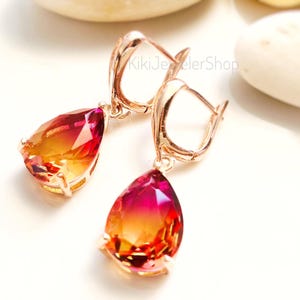 May include: A pair of rose gold earrings with teardrop-shaped, multi-colored gemstones. The gemstones transition from a deep red to a vibrant orange and yellow. The earrings have a lever-back closure.