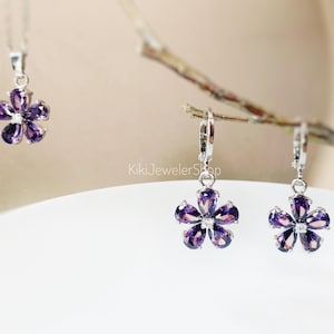Op de afbeelding: Een sieradenset met een bloemdesign met paarse edelstenen. De set bevat een hangerketting en een paar oorbellen. De oorbellen hebben een beugel sluiting. De sieraden staan tegen een zachte, vage achtergrond.