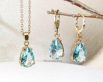 May include: A gold-toned jewelry set featuring a necklace and a pair of earrings. Each piece showcases a teardrop-shaped, light blue gemstone. The earrings are lever-back style. The necklace has a delicate chain. The jewelry is displayed on a light background.