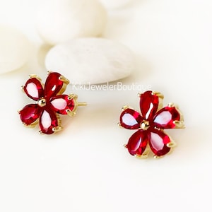 May include: A pair of gold stud earrings shaped like five-petaled flowers, each featuring vibrant red pear-cut gemstones set in a gold-tone metal frame. The text KikiJewelerBoutique is visible in the center. These earrings have a post back.