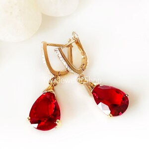 May include: Dangle earrings with teardrop-shaped, faceted red gemstones. The earrings have gold-colored settings and accents, with small clear stones set along the top of the earring hooks. The earrings are displayed on a white background.