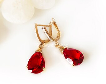 Peut inclure: Une paire de boucles d'oreilles pendantes dorées avec de grosses pierres rouges en forme de poire suspendues à des fermoirs articulés. Les fermoirs sont ornés de petites pierres claires sur le bord avant, contrastant avec les pierres rouges.