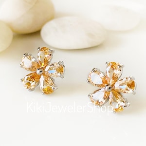 May include: A pair of flower-shaped stud earrings. Each earring features five amber-colored, pear-cut gemstones set in a silver-toned metal. The earrings are displayed on a white surface with white stones in the background.
