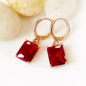 May include: A pair of gold-toned hoop earrings featuring rectangular, faceted deep red gemstones suspended from each hoop. The earrings have a hinged closure mechanism and are marked with the text KikiJewelerShop at the bottom center.