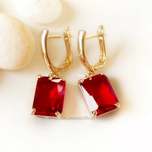 以下が含まれることがあります： 長方形の深い赤色の宝石が付いた、ゴールドトーンのイヤリングのペア。イヤリングはレバーバッククロージャーで、白い表面に表示されています。「KikiJewelerShop」というショップ名が画像に表示されています。