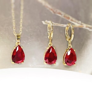 May include: A gold necklace with a teardrop-shaped, deep red pendant, and a matching set of gold earrings with similar red teardrop gems. The jewellery set is displayed on a neutral background.