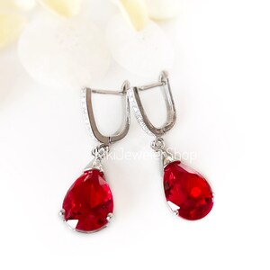May include: A pair of silver-toned drop earrings featuring vibrant red, pear-shaped faceted stones suspended from hinged hoops. The hoops are accented with small clear stones along the front. The text KikiJewelerShop is visible in the center.