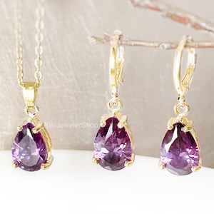 Puede incluir: Un conjunto de joyería en tono dorado que consta de un collar con un colgante de piedra preciosa púrpura en forma de pera y un par de pendientes a juego. Cada pieza presenta una piedra púrpura facetada en un engaste dorado. El texto KikiJewelerShop es visible.