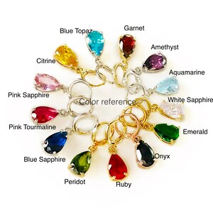 Op de afbeelding: Een verzameling edelsteen oorbellen in verschillende kleuren, waaronder blauwe topaas, citrien, granaat, amethist, aquamarijn, roze saffier, witte saffier, roze toermalijn, blauwe saffier, peridot, robijn, onyx en smaragd. Met een "color reference" label.