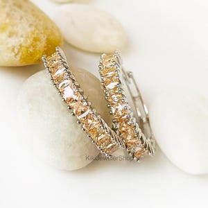 May include: Two silver-toned hoop earrings, each adorned with a row of rectangular, light-orange gemstones. The earrings rest on white and light-colored stones, with a blurred background. The earrings are a jewelry item.