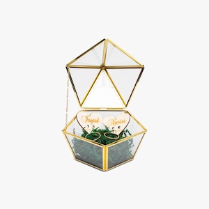 May include: A geometric glass and gold ring box with a hinged lid. Inside, two wedding rings rest on green moss, with two wooden heart-shaped tags engraved with names. A chain connects the lid to the box.