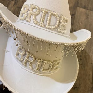 Puede incluir: Dos sombreros de vaquero blancos, uno encima del otro. Cada sombrero presenta la palabra "BRIDE" escrita con cuentas perladas. El ala de cada sombrero est&aacute; adornada con brillantes colgantes de color plateado, a&ntilde;adiendo un toque de brillo al accesorio nupcial.