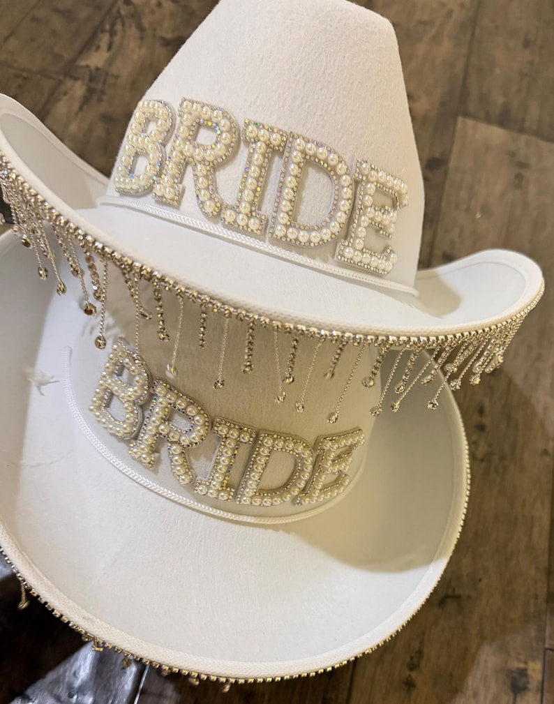 Puede incluir: Dos sombreros de vaquero blancos, uno encima del otro. Cada sombrero presenta la palabra "BRIDE" escrita con cuentas perladas. El ala de cada sombrero est&aacute; adornada con brillantes colgantes de color plateado, a&ntilde;adiendo un toque de brillo al accesorio nupcial.