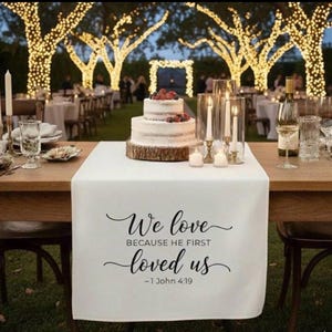 May include: A wedding reception table is set with a white runner featuring the text "We love because he first loved us - 1 John 4:19." A two-tiered cake with berries sits on a wooden base. Candles and glassware complete the elegant setting.