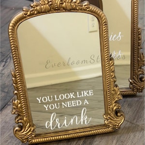 Puede incluir: Espejo dorado ornamentado con el texto "YOU LOOK LIKE YOU NEED A drink" en escritura blanca. El espejo tiene un diseño decorativo y vintage, probablemente utilizado para la decoración del hogar o eventos. El espejo es rectangular con esquinas redondeadas.
