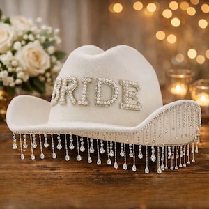 Puede incluir: Un sombrero de vaquero blanco con la palabra "BRIDE" escrita con cuentas de perlas. El ala del sombrero est&aacute; adornada con l&aacute;grimas colgantes de cristal. Un accesorio nupcial.