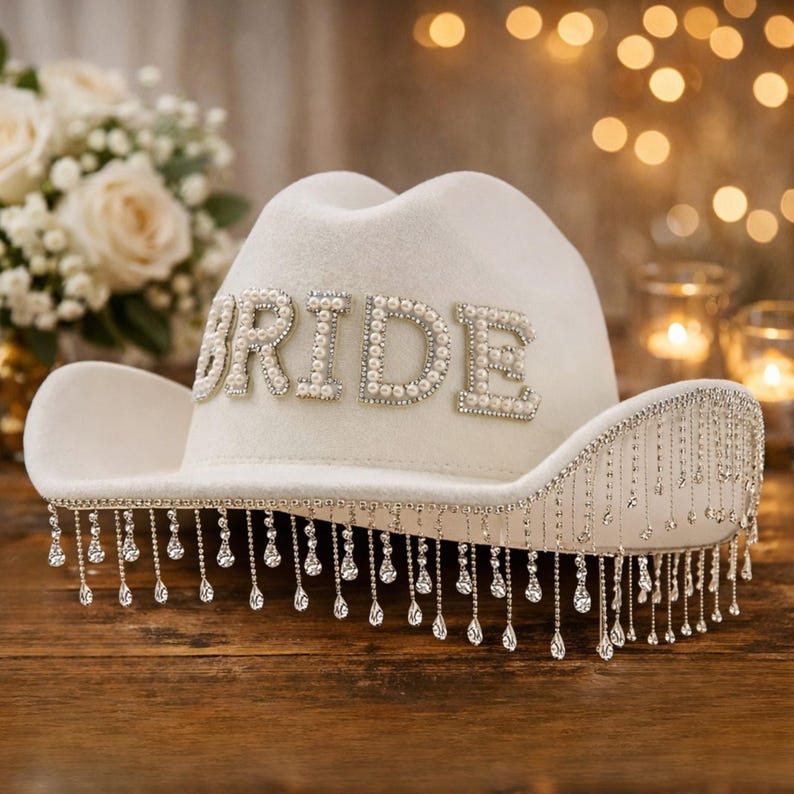 Puede incluir: Un sombrero de vaquero blanco con la palabra "BRIDE" escrita con cuentas de perlas. El ala del sombrero est&aacute; adornada con l&aacute;grimas colgantes de cristal. Un accesorio nupcial.