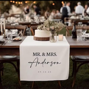 Peut inclure: Chemin de table blanc avec le texte "MR. & MRS. Anderson 09.14.25" sur une table en bois pour une réception de mariage. Gâteau, fleurs et bougies décorent la table.