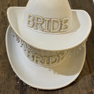 Puede incluir: Dos sombreros de vaquero blancos apilados uno encima del otro. La palabra "BRIDE" est&aacute; escrita con cuentas de perlas en la parte delantera de cada sombrero. El ala del sombrero inferior est&aacute; adornada con cadenas colgantes de color plateado.