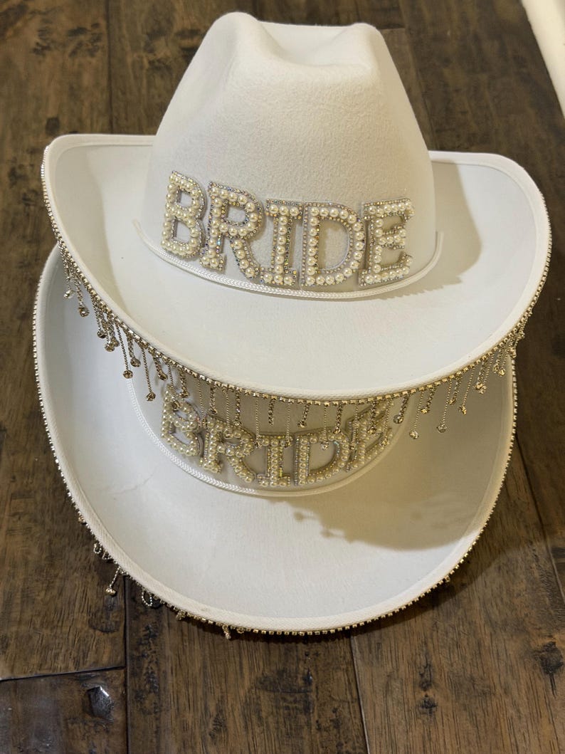 Puede incluir: Dos sombreros de vaquero blancos apilados uno encima del otro. La palabra "BRIDE" est&aacute; escrita con cuentas de perlas en la parte delantera de cada sombrero. El ala del sombrero inferior est&aacute; adornada con cadenas colgantes de color plateado.