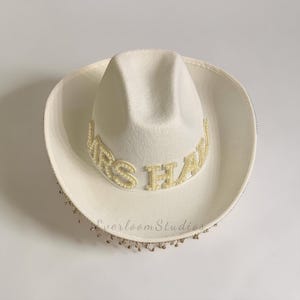Puede incluir: Un sombrero de vaquero blanco con el texto "MRS HAW" en letras doradas y detalles de perlas. El ala est&aacute; adornada con peque&ntilde;as cuentas doradas. Un accesorio nupcial.