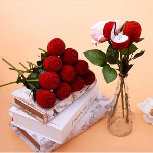 May include: A bouquet of red velvet rose-shaped ring boxes and a bouquet of roses with a ring box. The ring box is open, revealing a diamond ring. The bouquets are arranged on top of books and in a glass vase.