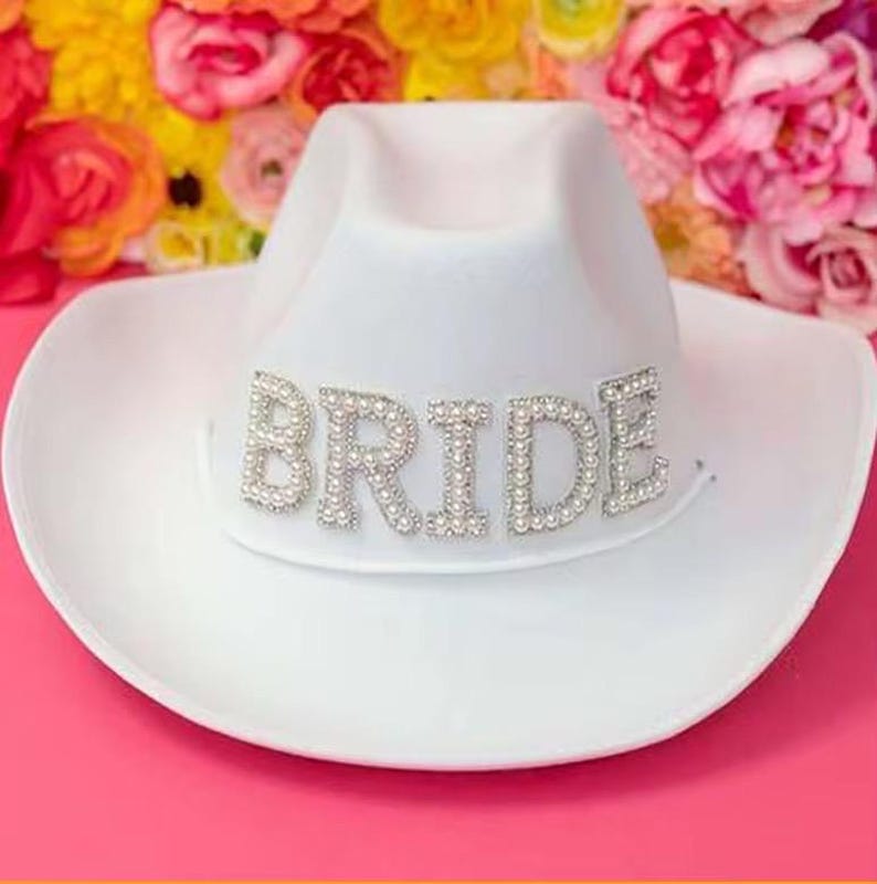 Puede incluir: Un sombrero de vaquero blanco con la palabra "BRIDE" escrita en letras plateadas con detalles de perlas. El sombrero est&aacute; sobre un fondo de flores coloridas, perfecto para una despedida de soltera o una fiesta de compromiso.