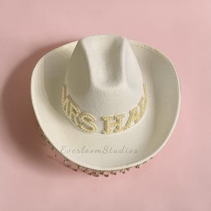 Puede incluir: Un sombrero de vaquero blanco con el texto "MRS HAW" en letras doradas. El sombrero est&aacute; adornado con perlas y un ribete de cuentas. El sombrero est&aacute; sobre un fondo rosa. El texto "Everloom Studios" est&aacute; impreso en el sombrero.