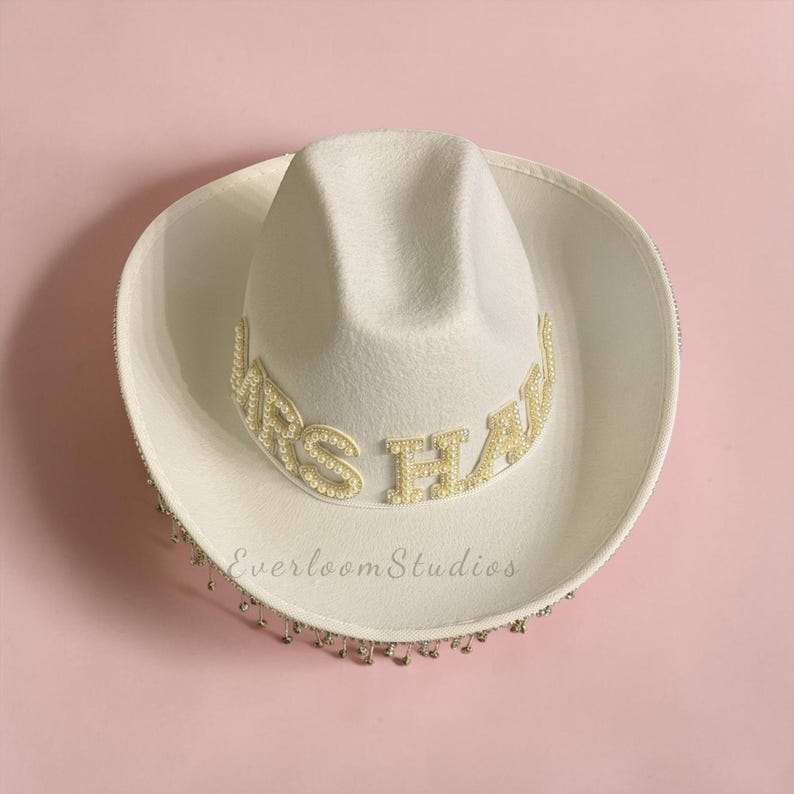 Puede incluir: Un sombrero de vaquero blanco con el texto "MRS HAW" en letras doradas. El sombrero est&aacute; adornado con perlas y un ribete de cuentas. El sombrero est&aacute; sobre un fondo rosa. El texto "Everloom Studios" est&aacute; impreso en el sombrero.