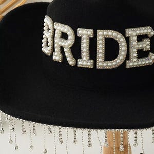 Puede incluir: Sombrero fedora negro con la palabra "BRIDE" en cuentas de perlas. El ala del sombrero est&aacute; adornada con cadenas colgantes de cristal plateado. Un accesorio de moda para una boda.