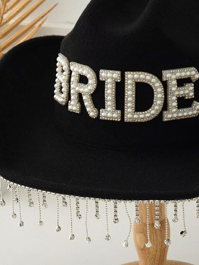 Puede incluir: Sombrero fedora negro con la palabra "BRIDE" en cuentas de perlas. El ala del sombrero est&aacute; adornada con cadenas colgantes de cristal plateado. Un accesorio de moda para una boda.