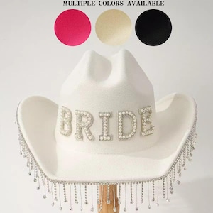 Puede incluir: Un sombrero de vaquero blanco con la palabra "BRIDE" en perlas. El ala del sombrero est&aacute; adornada con brillantes plateados colgantes. La imagen muestra tres opciones de color: rosa, beige y negro. El texto dice "M&Uacute;LTIPLES COLORES DISPONIBLES".
