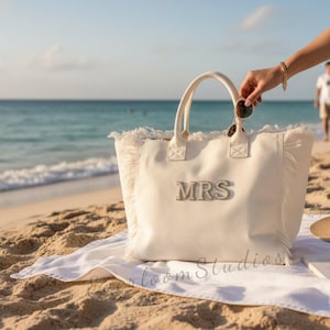 May include: Cream-colored tote bag with the word "MRS" spelled out in silver rhinestones. The bag has fringe detailing and is sitting on a white towel on a sandy beach. The ocean is visible in the background.