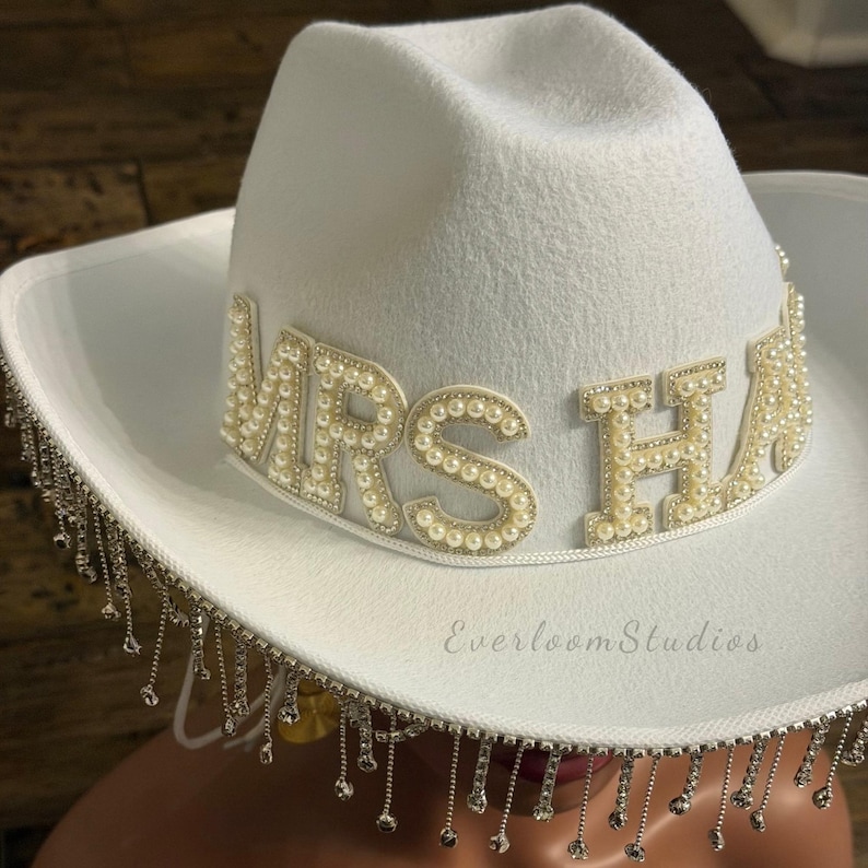 Puede incluir: Sombrero de vaquero blanco con el texto "MRS HA" en adornos de perlas. El sombrero presenta una banda decorativa de pedrer&iacute;a y flecos de cristal colgantes. Un accesorio nupcial.