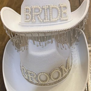 Puede incluir: Dos sombreros de vaquero blancos, uno encima del otro. El sombrero superior tiene la palabra "BRIDE" en perlas de imitaci&oacute;n, con flecos plateados colgantes. El sombrero inferior tiene "GROOM" en perlas.