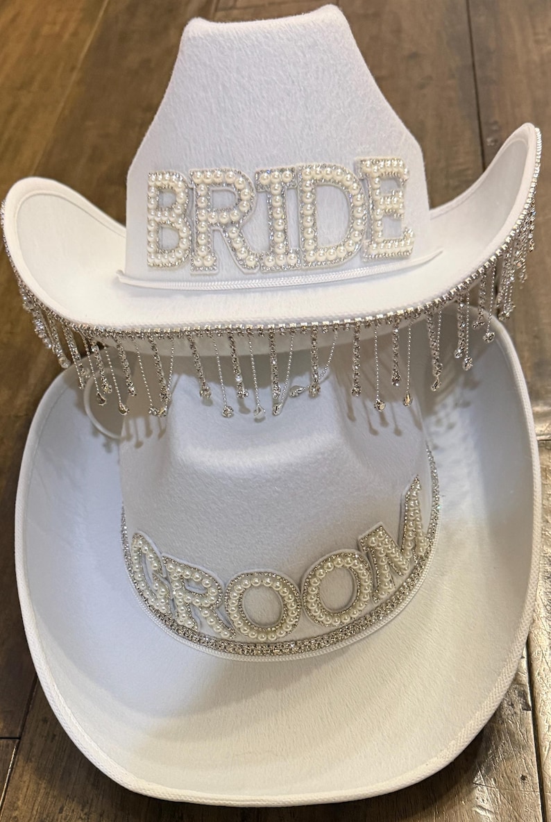 Puede incluir: Dos sombreros de vaquero blancos, uno encima del otro. El sombrero superior tiene la palabra "BRIDE" en perlas de imitaci&oacute;n, con flecos plateados colgantes. El sombrero inferior tiene "GROOM" en perlas.