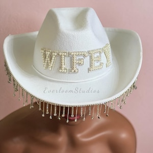 Puede incluir: Un sombrero de vaquero blanco con la palabra "WIFEY" escrita con adornos de perlas. El sombrero presenta una franja plateada alrededor del borde y el texto "EverloomStudios" en la parte delantera.
