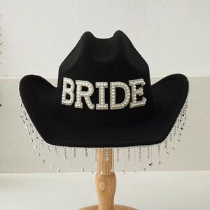 Puede incluir: Sombrero de vaquero negro con la palabra "BRIDE" en perlas blancas. El sombrero tiene una franja plateada alrededor del borde. Un accesorio de moda para una despedida de soltera o una boda.