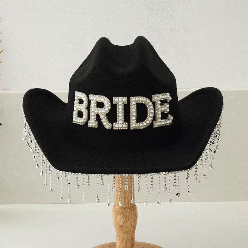 Puede incluir: Sombrero de vaquero negro con la palabra "BRIDE" en perlas blancas. El sombrero tiene una franja plateada alrededor del borde. Un accesorio de moda para una despedida de soltera o una boda.
