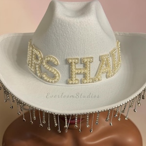 Puede incluir: Sombrero de vaquero blanco con el texto "MRS HAW" en letras perladas. El ala del sombrero est&aacute; adornada con brillantes plateados colgantes. Un accesorio nupcial.