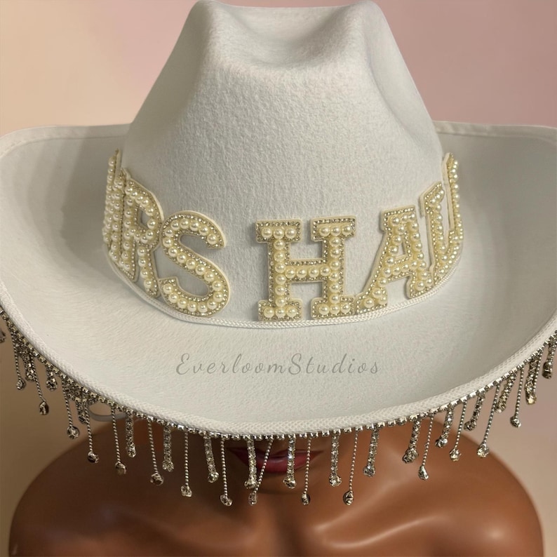 Puede incluir: Sombrero de vaquero blanco con el texto "MRS HAW" en letras perladas. El ala del sombrero est&aacute; adornada con brillantes plateados colgantes. Un accesorio nupcial.