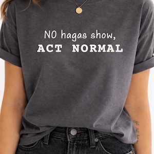 May include: A charcoal gray short-sleeve t-shirt featuring the white printed text NO hagas show, ACT NORMAL. The shirt has a crew neckline and a relaxed fit with rolled sleeves, constructed from a soft, washed-out fabric with a vintage appearance.