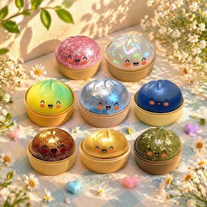 Puede incluir: Una colección de coloridos recipientes con forma de dumpling sonriente. Cada recipiente tiene un color y una textura diferentes, incluyendo purpurina rosa, blanco iridiscente, azul, amarillo, marrón, dorado y verde. Los recipientes están en soportes de color canela.