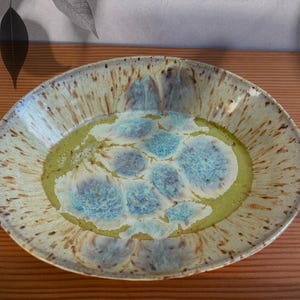 May include: A handmade ceramic serving platter with a unique glaze. The oval-shaped dish features a light beige exterior with brown speckles. The interior has a green border and a central design of blue and white patterns.