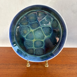 May include: A round ceramic bowl featuring a crystalline glaze with a deep blue and teal color palette. The interior surface displays a distinct pattern of dark, starburst-like crystalline formations surrounded by lighter green and blue hues.