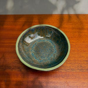 May include: A round ceramic bowl featuring a glossy interior glaze with swirling patterns of deep blue, teal, and earthy brown tones. The exterior rim is finished in a solid, muted light green glaze, providing a contrast to the dark, mottled center.