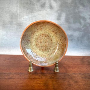 May include: A round ceramic bowl featuring a speckled glaze with a blend of warm tan, beige, and brown tones. The bowl is supported by a small, metallic, two-pronged stand that holds the piece in an upright position for display purposes.
